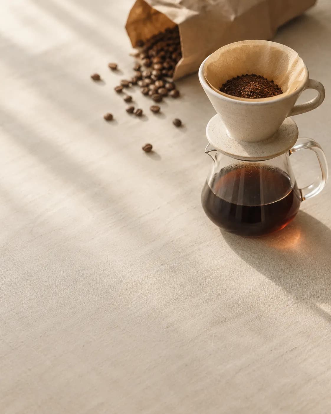 Pour-over dripper and glass server on linen in morning light, with scattered coffee beans